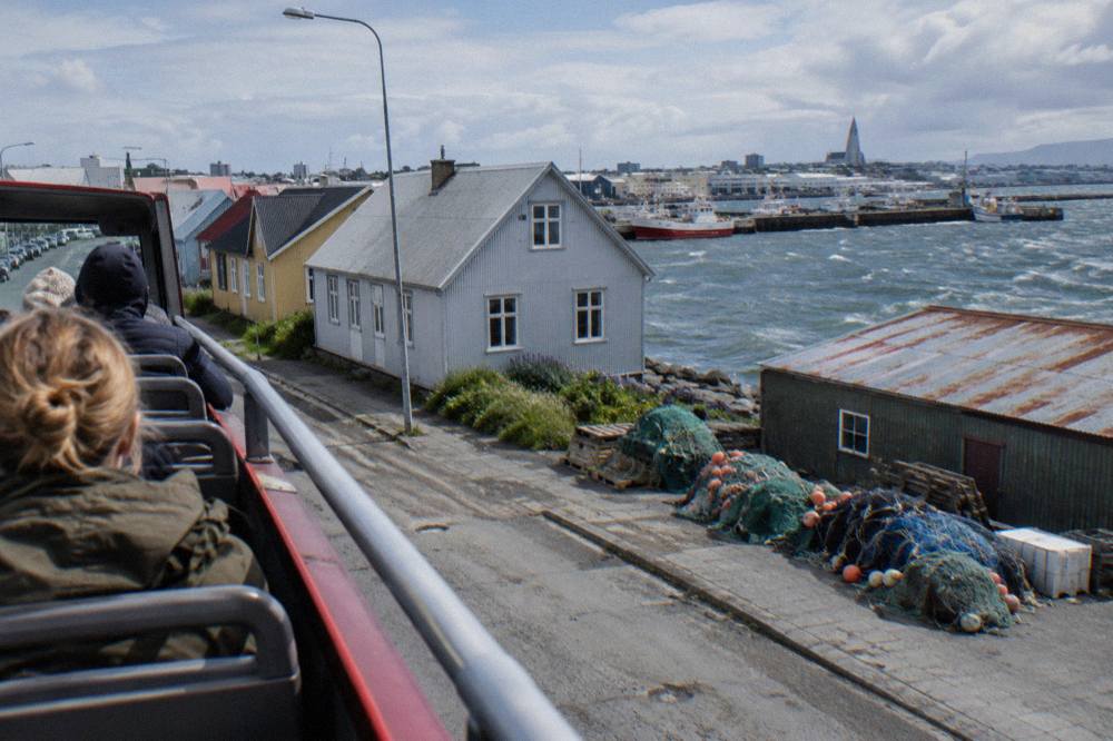 An open top tour bus moving along a waterfront street past low colourful houses