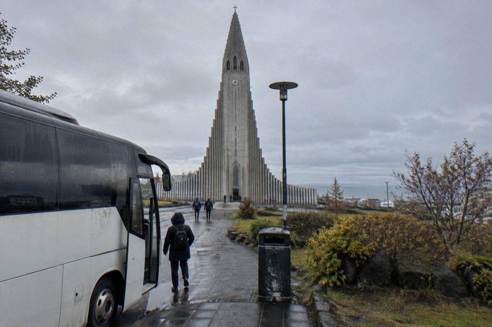 A tour bus parked at a stop beside a tall concrete church tower under a grey sky