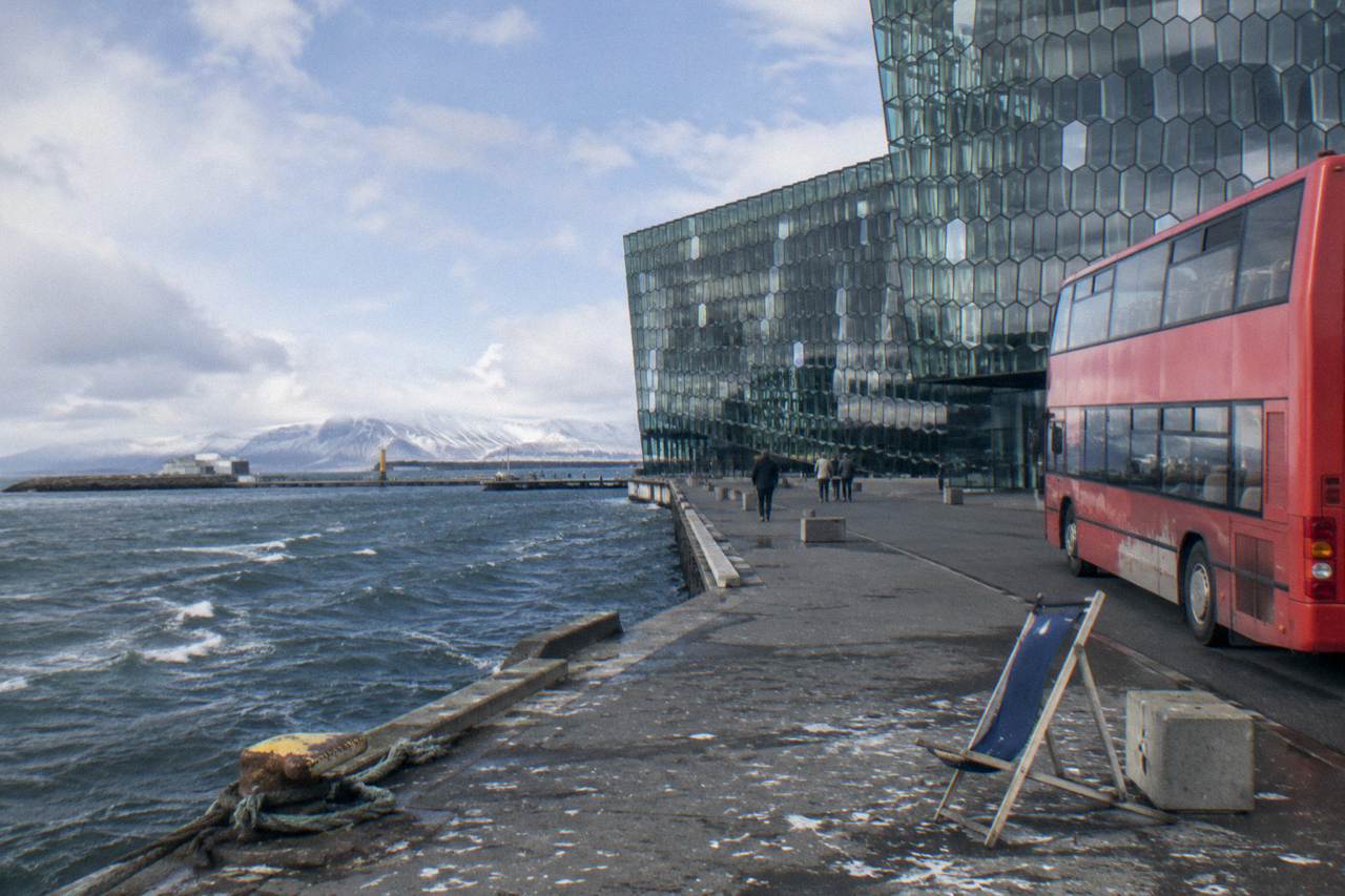 A red double decker tour bus parked beside a modern harbourfront concert hall in a Nordic capital