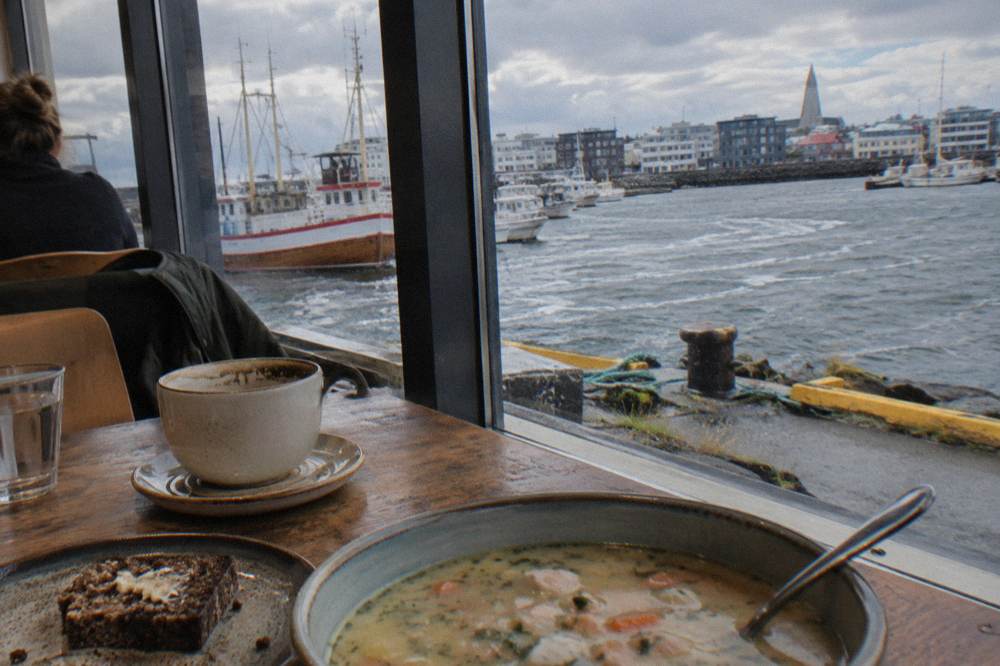 Café table by a window with coffee, bread and soup in warm indoor light