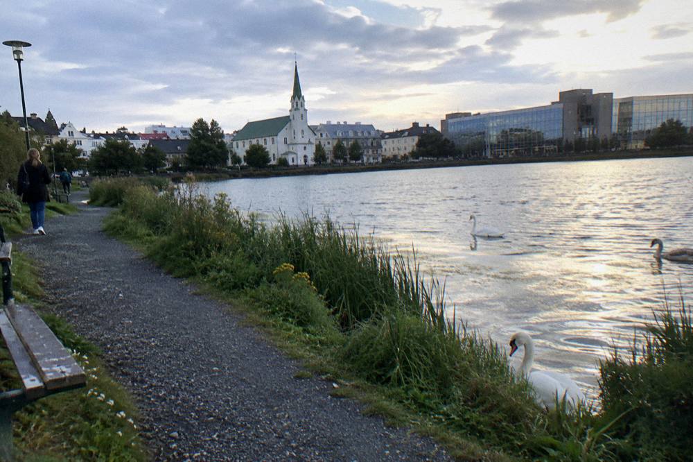 A stone path curving along the edge of a small lake beside modern civic buildings in the city centre
