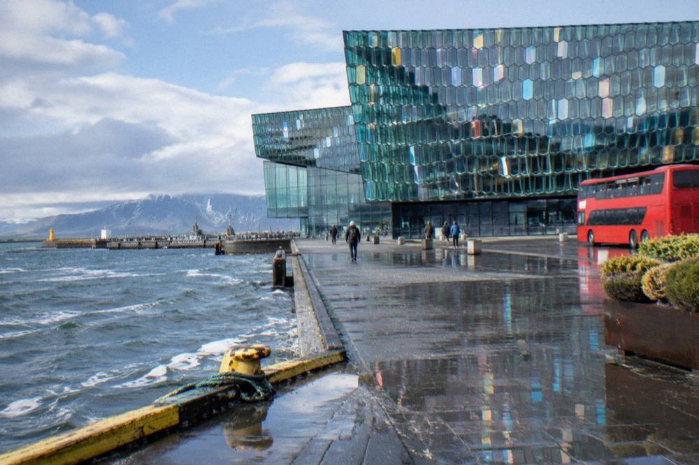 A red double-decker tour bus parked beside the glass facade of Harpa concert hall