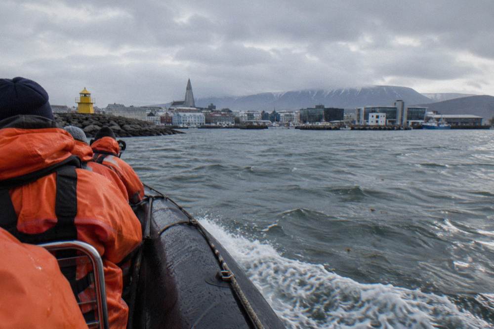 Passengers in orange exposure suits seated on a rigid inflatable boat heading out of a harbour
