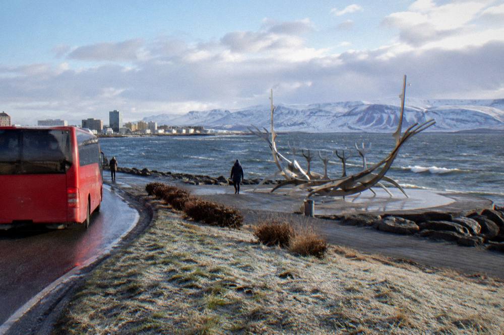 A sightseeing bus rounding a curve on the Reykjavik seafront with snow-dusted mountains across the water
