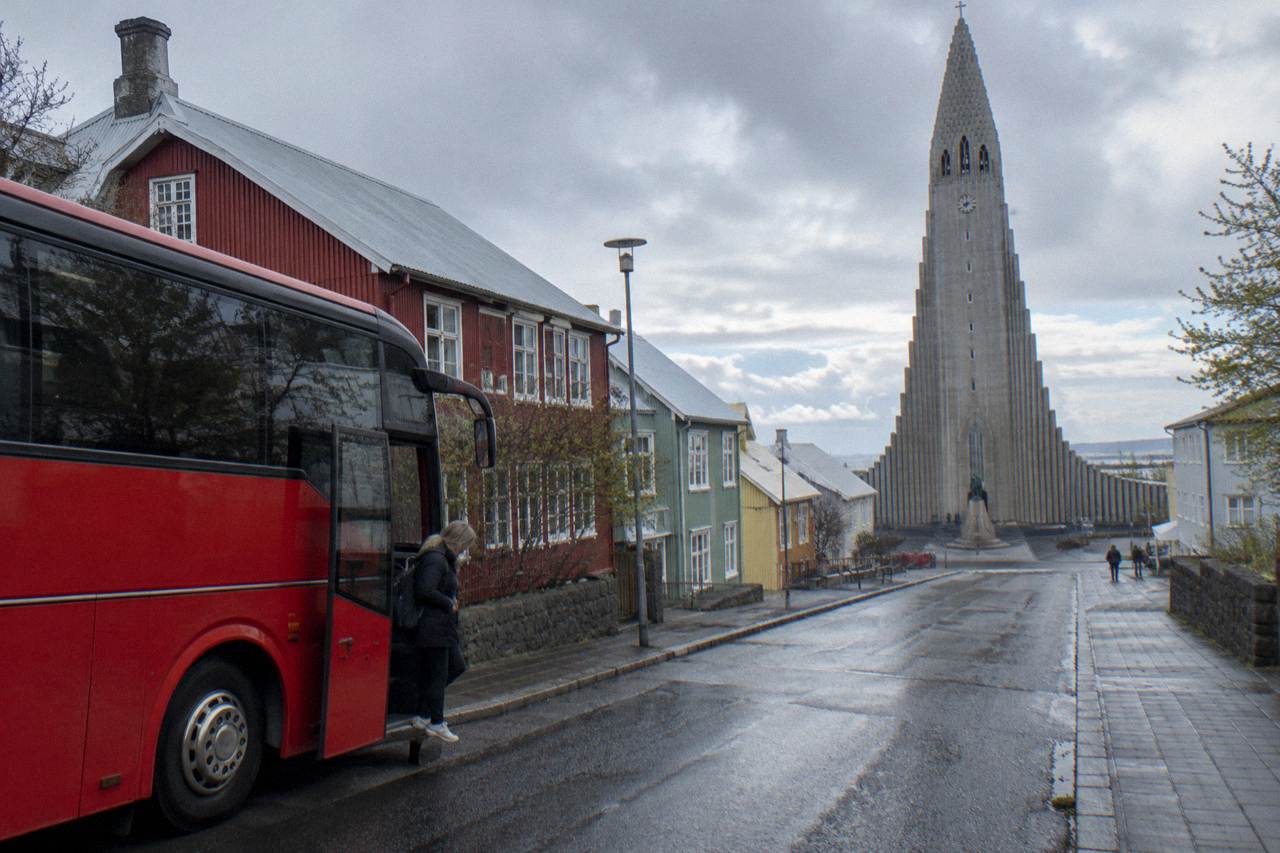 A red sightseeing bus parked on a Reykjavik street with the Hallgrimskirkja church tower behind it under a grey sky