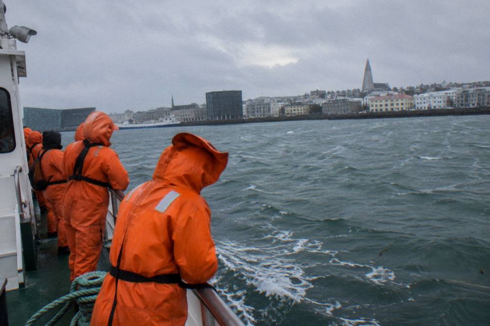 Passengers in orange coveralls watching the water from a boat rail in cold daylight