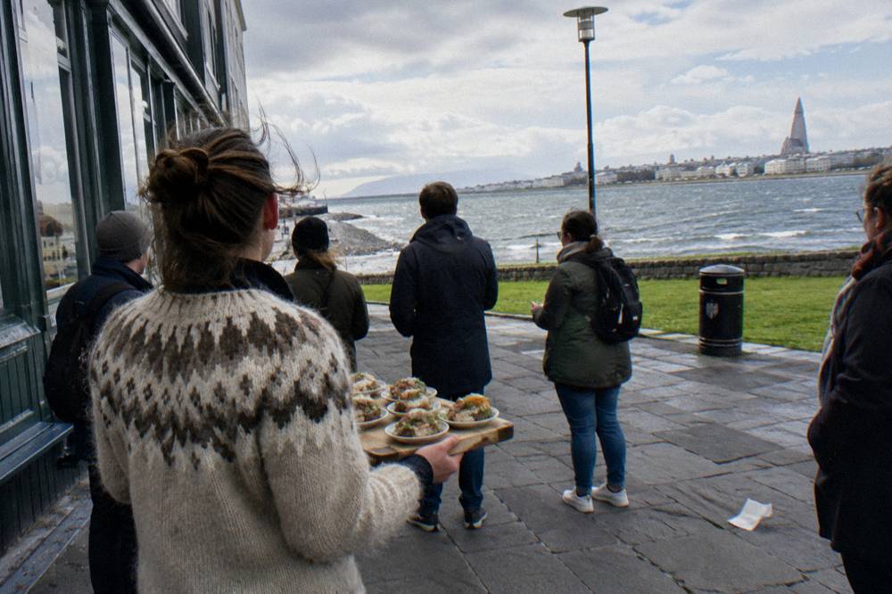 A guide presenting small tasting plates to a small standing group outside a shopfront